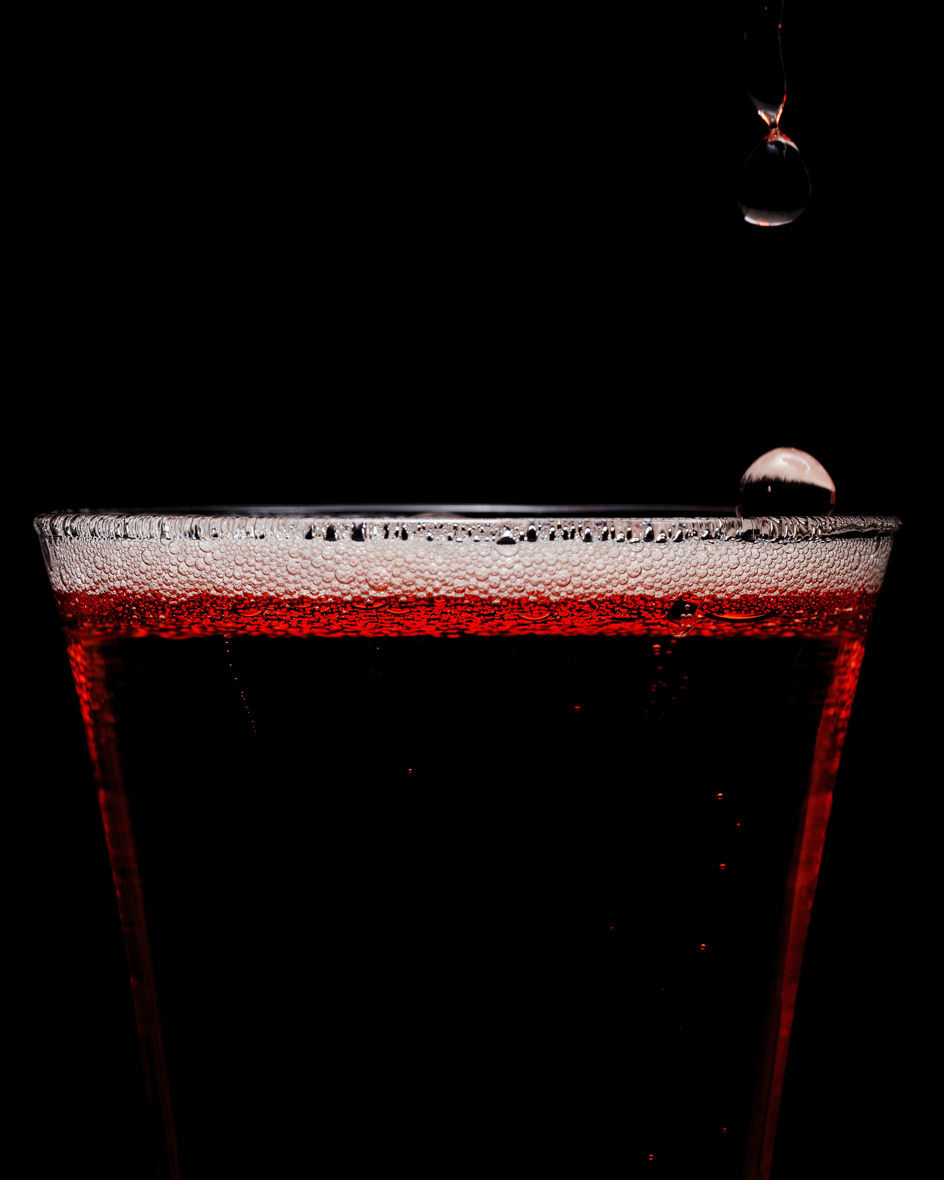 A glass filled with dark red liquid sits against a black background, with foam on top and several droplets falling above it.