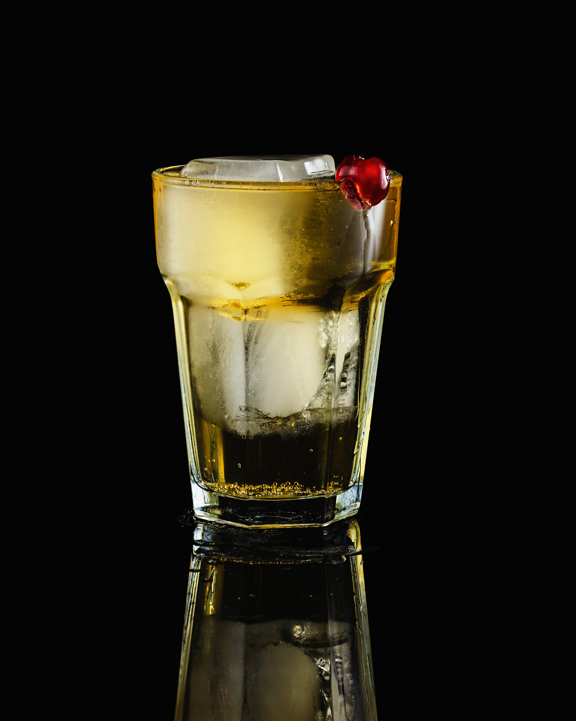 A ginger ale drink with ice cubes and candied cherry against a dark background.