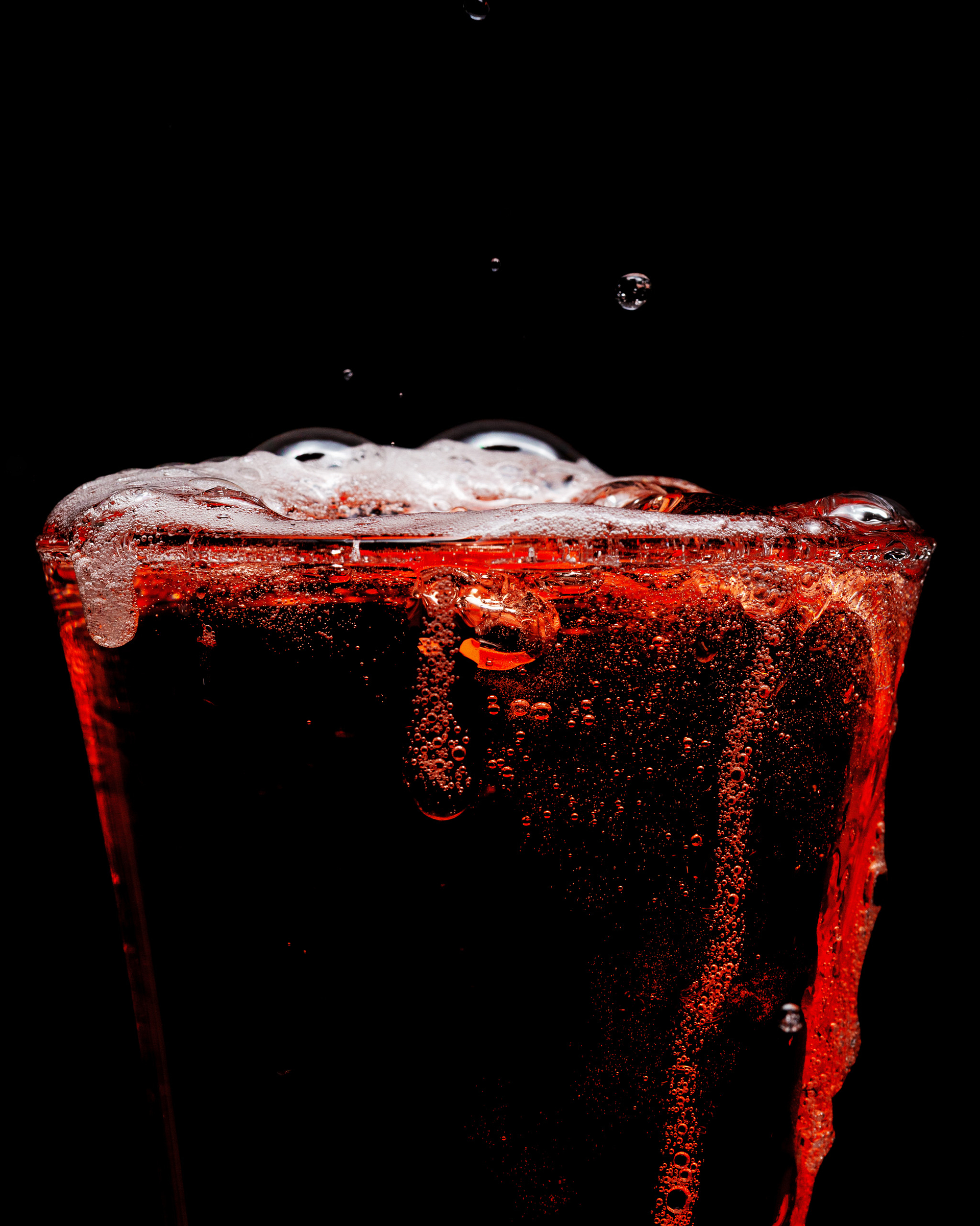 A close-up view of a glass filled with dark red liquid shows foam, bubbles, and splashes against a black background.