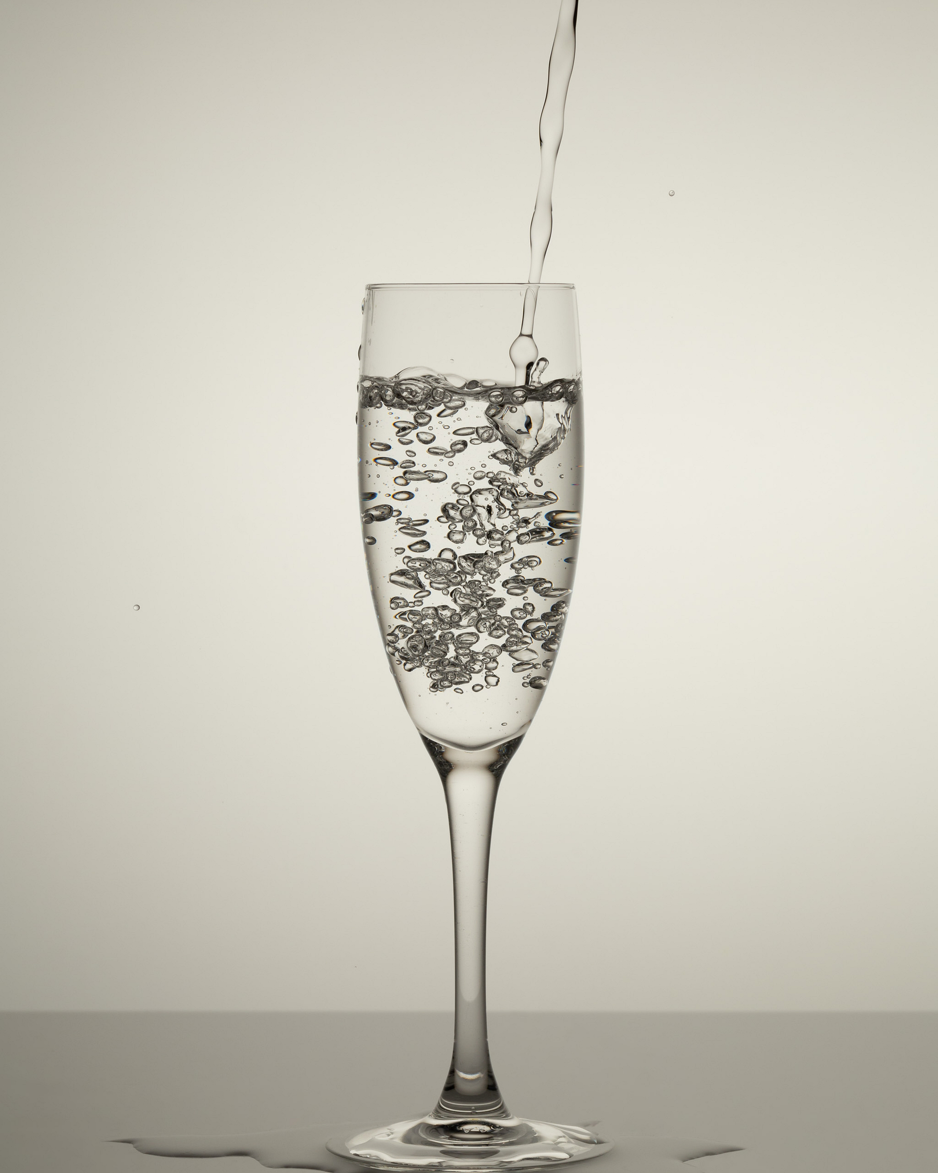 Water pours into a tall glass filled with clear liquid, creating bubbles against a light background.